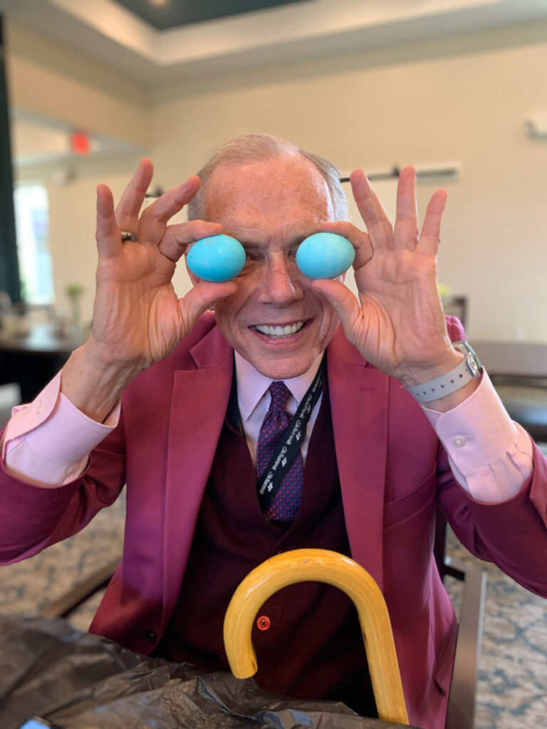 With a mischievous smile, a senior resident in a wine color suit and tie playfully holds two easter eggs up to his eyes, adding a touch of whimsy to his day.