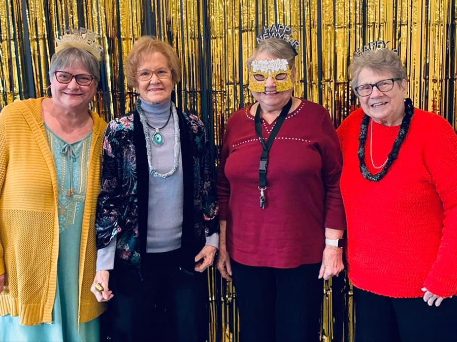 Four lovely senior women standing together in front of a gold curtain, ready for a new year celebration photo.