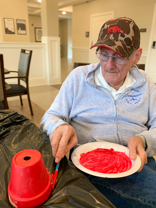 Senior man in a camo cap and glasses, painting a flower pot red at a table covered with a black plastic tablecloth during an arts and crafts activity in the community.