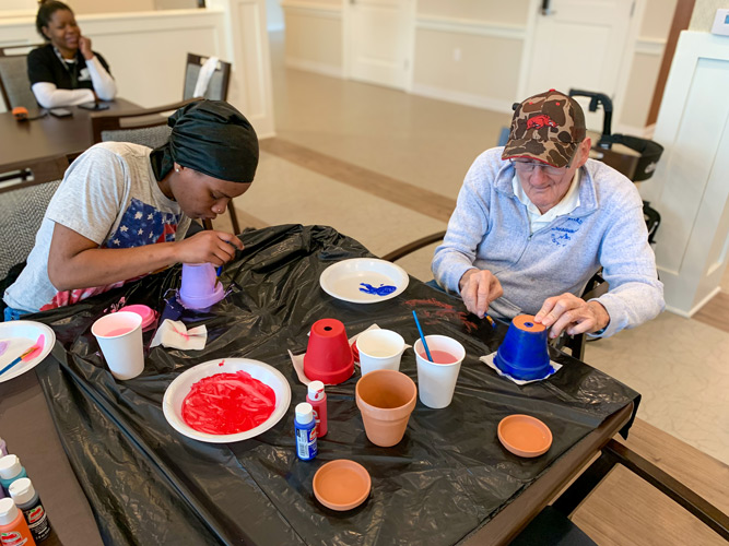 Senior living resident and a young woman painting flower pots at a table covered with a black plastic tablecloth, engaging in an arts and crafts activity.