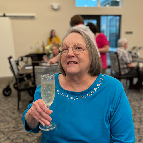 Senior lady in a blue top with rhinestones, smiling and holding a glass of sparkling drink during a social event in the community.