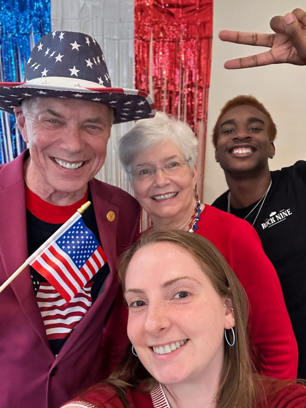 Four smiling people pose for a photo in front of red, white, and blue decorations. One person is holding a small American flag, celebrating a patriotic event.