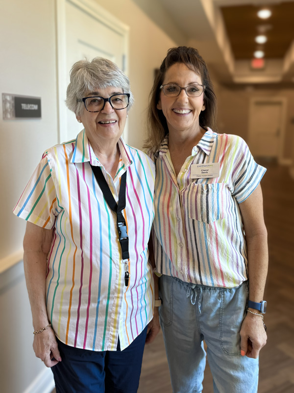 Two smiling women wearing striped shirts stand together in a brightly lit hallway. One is a senior resident, and the other appears to be a staff member.