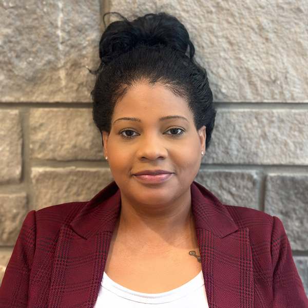Sheena Henry, Culinary Director at The Summit Senior Living, standing in front of a stone wall, wearing a red plaid blazer and white top with her hair in a bun.