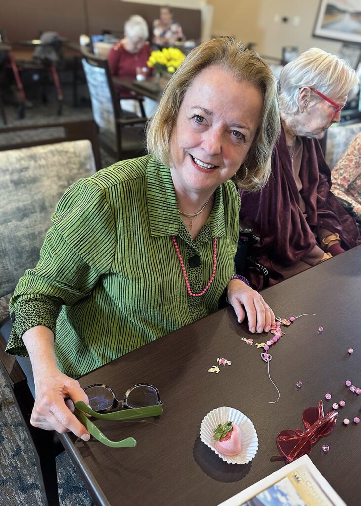 A smiling woman sits with other residents in the background, enjoying a crafting event with a chocolate covered strawberry to eat.