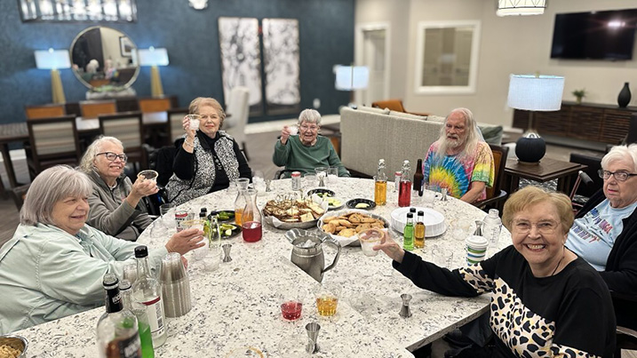 Residents smile, raising their glasses in a toast, around a table full of food and drink during a gathering.