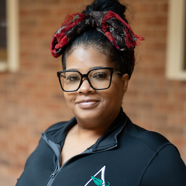 Sheena Henry, Culinary Director at The Summit Senior Living, smiling in a professional headshot, wearing glasses, a black zip-up jacket, and a patterned headband, with a softly blurred brick background.