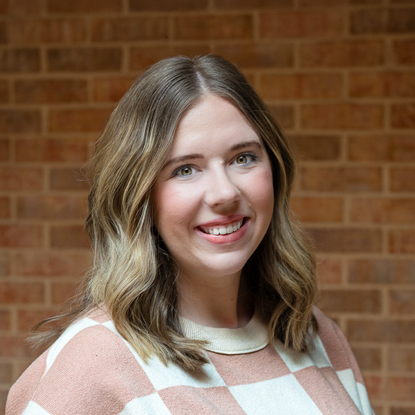 Briley Evans, Resident Services Director at The Summit Senior Living, smiling in a professional outdoor headshot with shoulder-length wavy blonde hair, a pink and cream sweater, and a softly blurred brick background.
