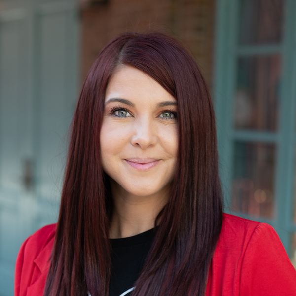 Ellen Cavazos, Executive Director at The Summit Senior Living, in a professional outdoor headshot with long auburn hair, bright eyes, a red blazer, and softly blurred brick and teal window details in the background.
