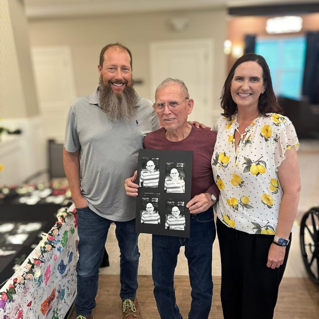 A senior man smiles alongside loved ones, holding a series of black and white portraits of himself.