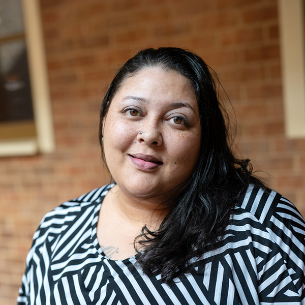 Whitney Dean, Wellness Director at The Summit Senior Living, looking at the camera in a professional outdoor headshot with long dark hair, a black and white patterned blouse, and a softly blurred brick background.