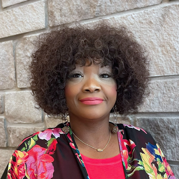 Amanda Briggs, Business Office Director at The Summit Senior Living, looking at the camera in a professional headshot with a short curly hairstyle, floral kimono, bright pink top, and a light gray stone wall background.