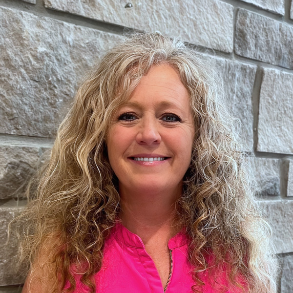 Joann Hall, Executive Director at The Summit Senior Living, smiling in a professional headshot with long curly blonde hair, a bright pink sleeveless top, and a light gray stone wall background.