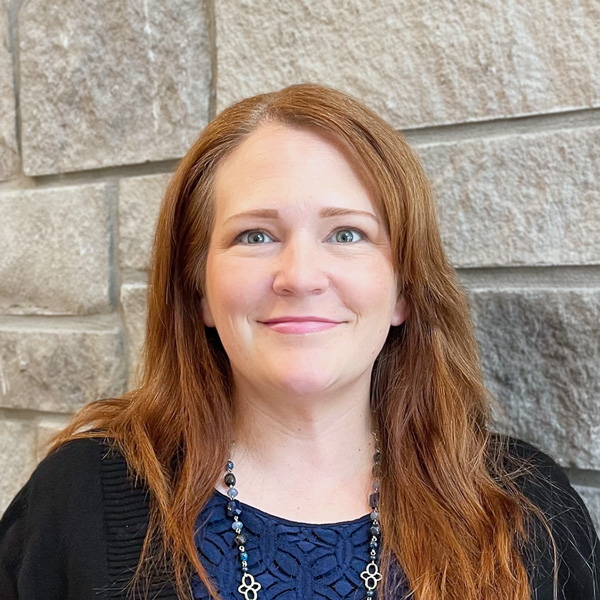 Katie Zrebski, Memory Care Director at The Summit Senior Living, looking at the camera in a professional headshot with long red hair, a blue blouse, layered necklaces, and a light gray stone wall background.