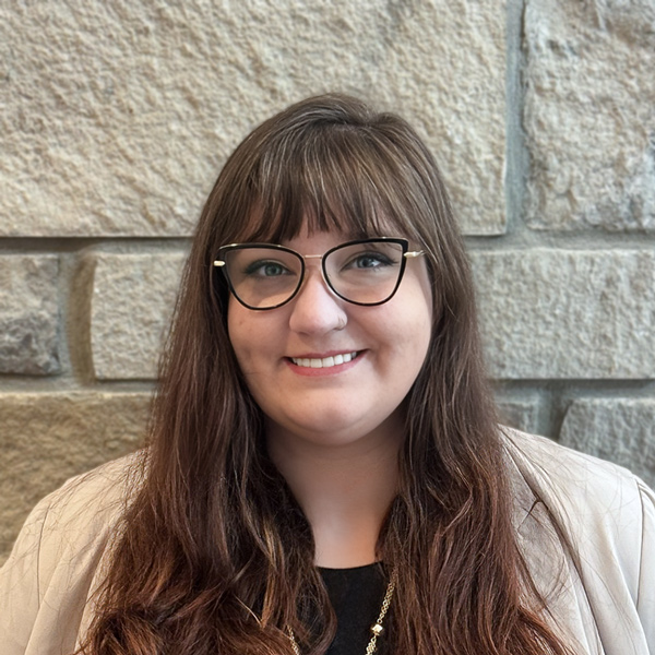 Aine Rickman, Senior Living Coordinator at The Summit Senior Living, smiling in a professional headshot with long brown hair, black glasses, a cream blazer, and a light stone wall background.